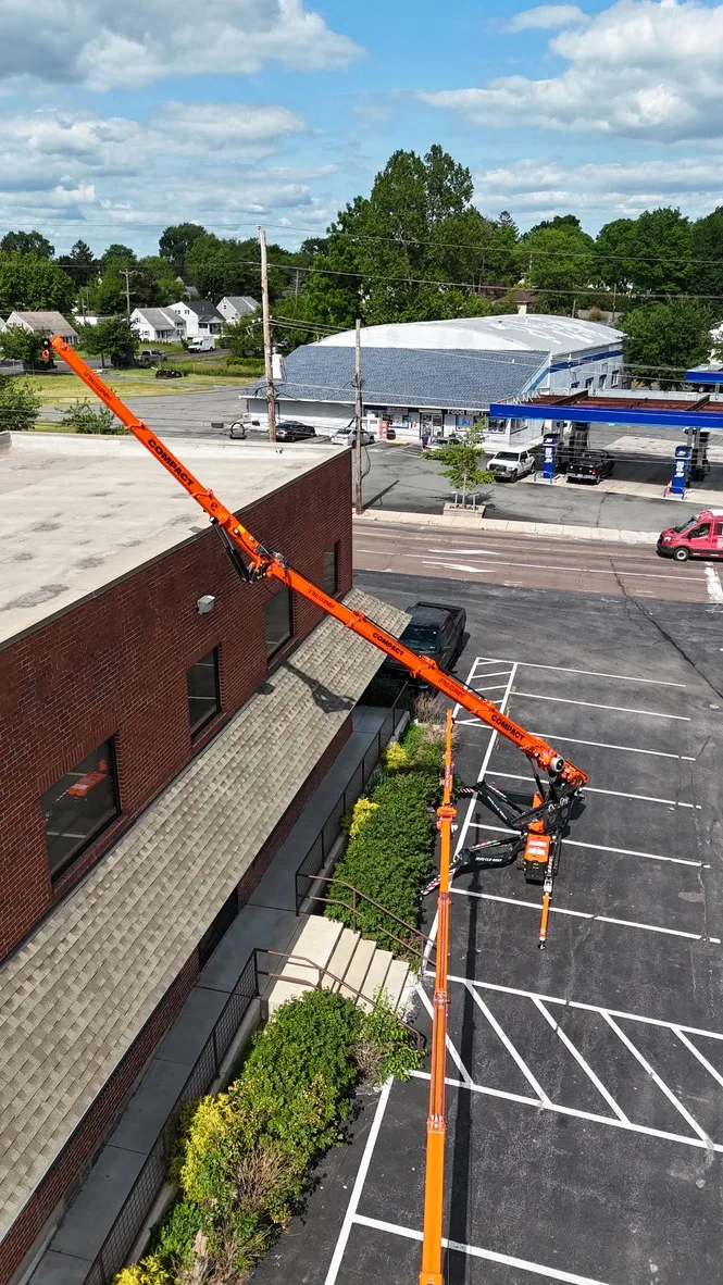 Aerial view of a Jekko mini crane set up in a parking lot with outriggers spread, boom and jib reaching over a brick building to the roof