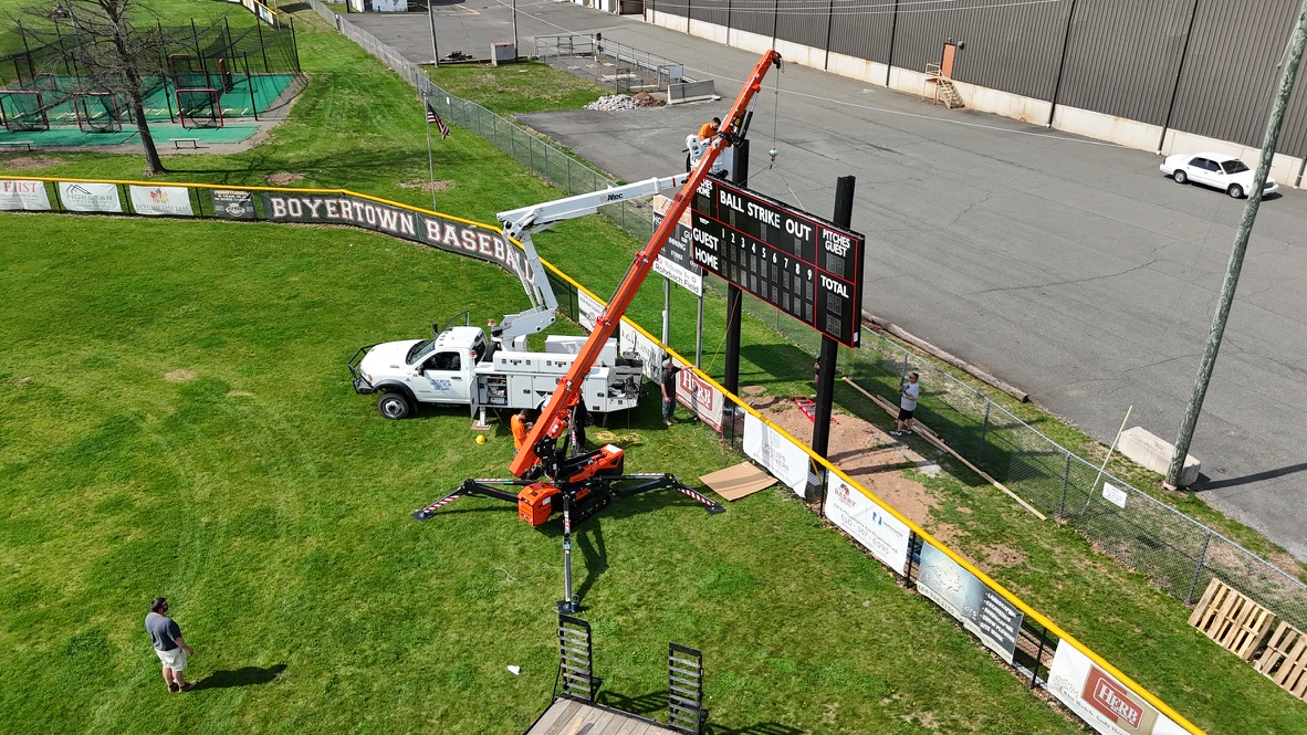 Compact spider lift and bucket truck installing a baseball field scoreboard from the outfield grass