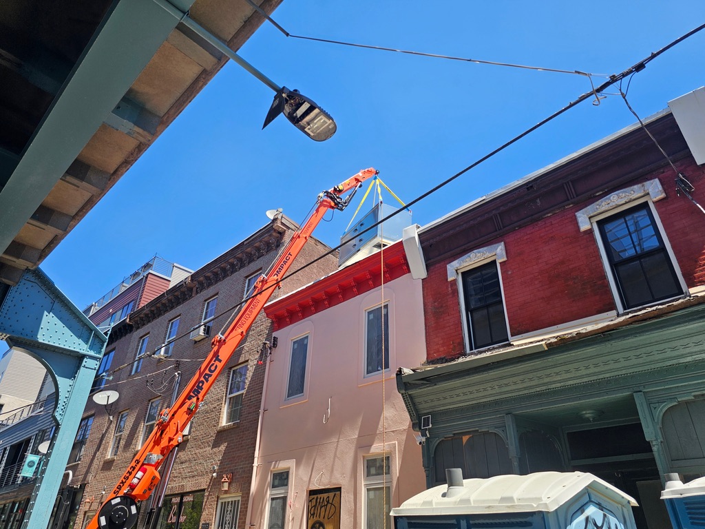 Compact boom lift setting a rooftop HVAC unit over a parapet on a tight city street