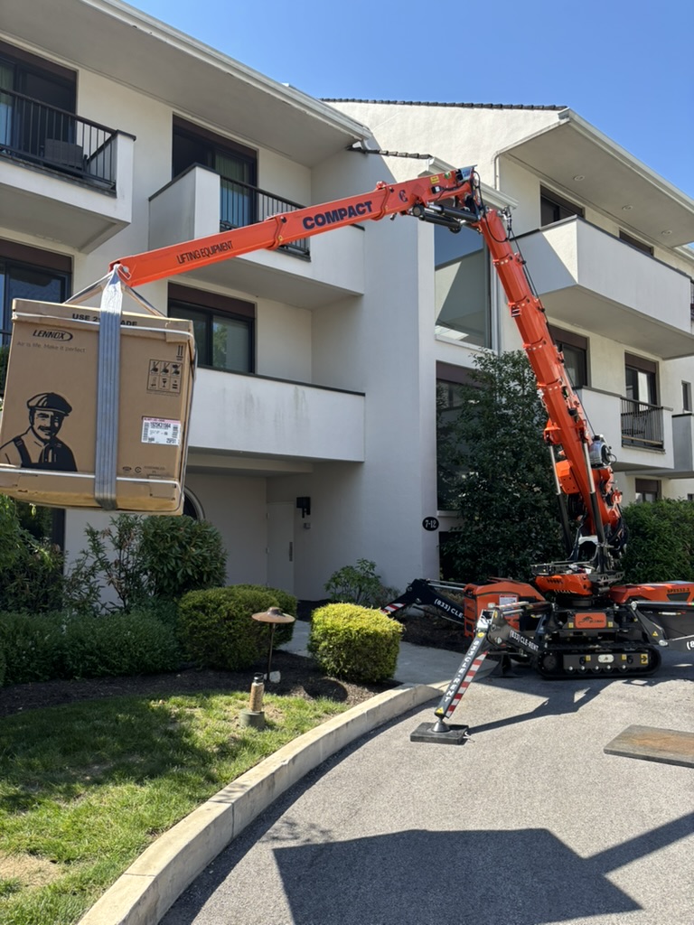 Compact crawler boom lift raising a boxed Lennox HVAC unit up to an upper-floor balcony from a residential driveway