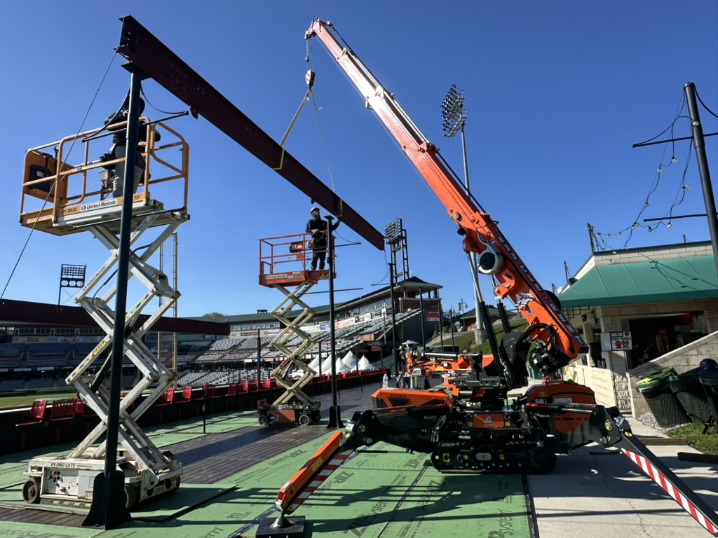 Compact spider crane lifting a structural steel beam over a stadium field with scissor lifts staged alongside